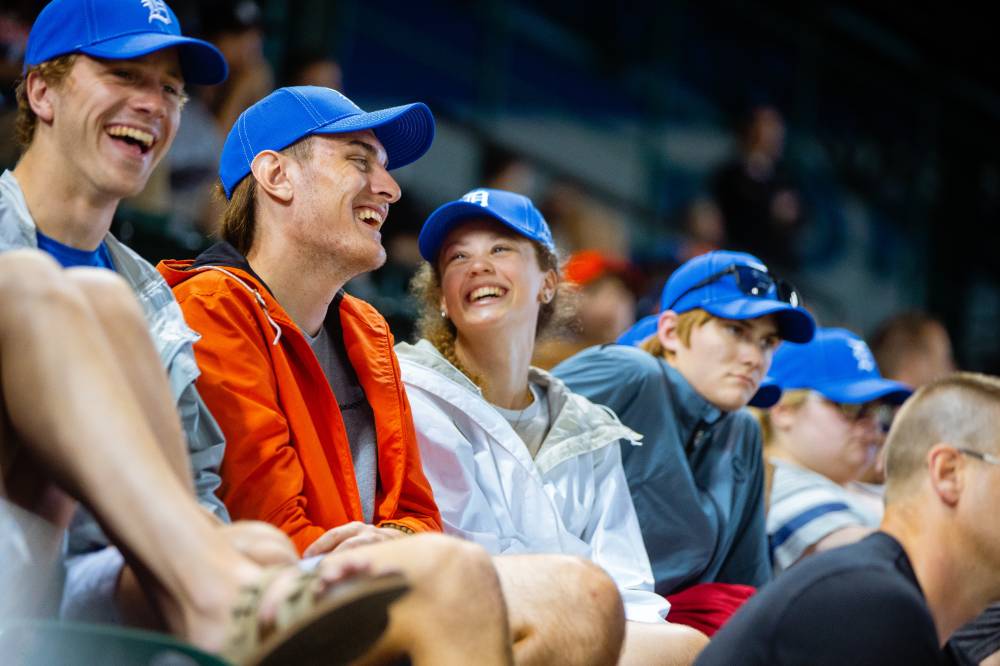 A row of fans wearing gvsu night tigers hats smiling and laughing with each other as they watch the game